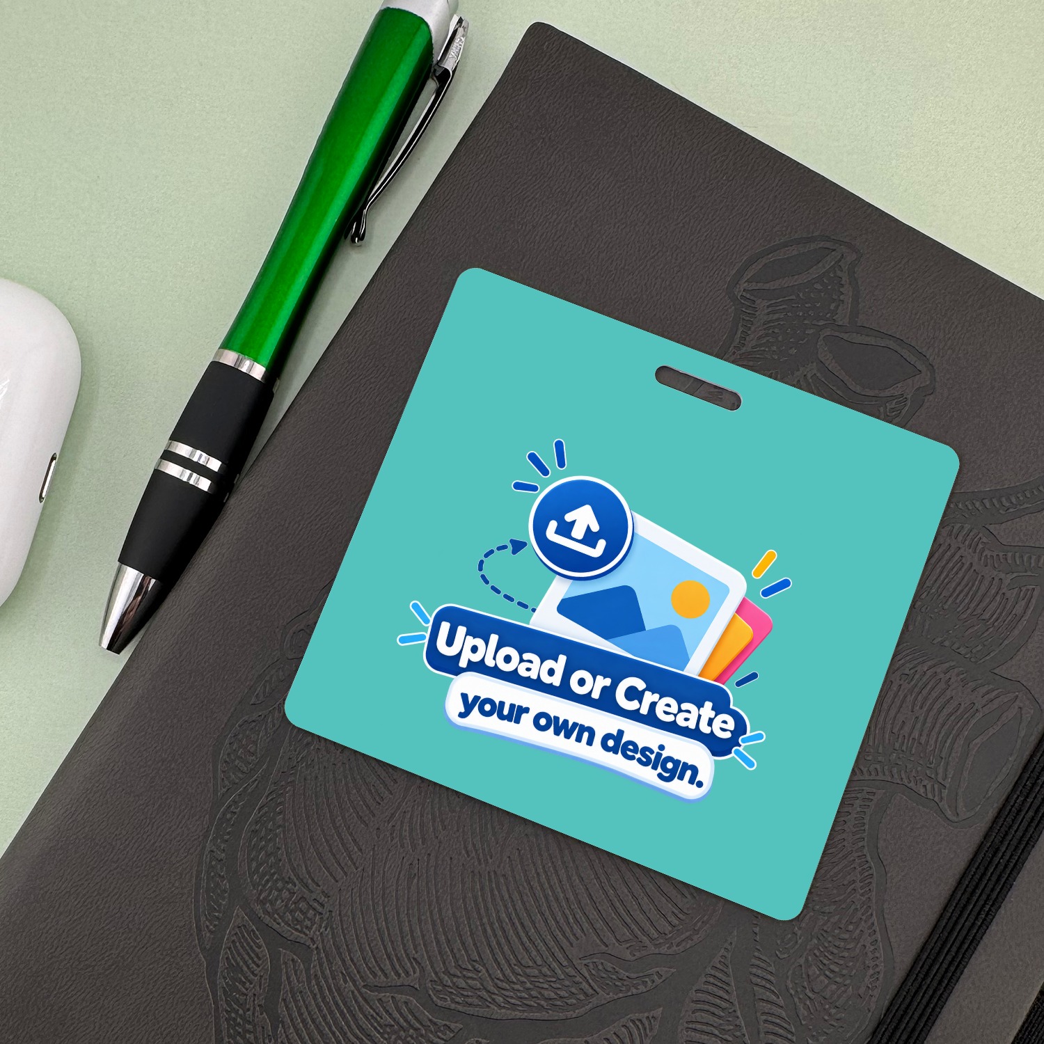 Teal horizontal ID badge with Upload or Create your own design graphic beside green ballpoint pen on dark notebook