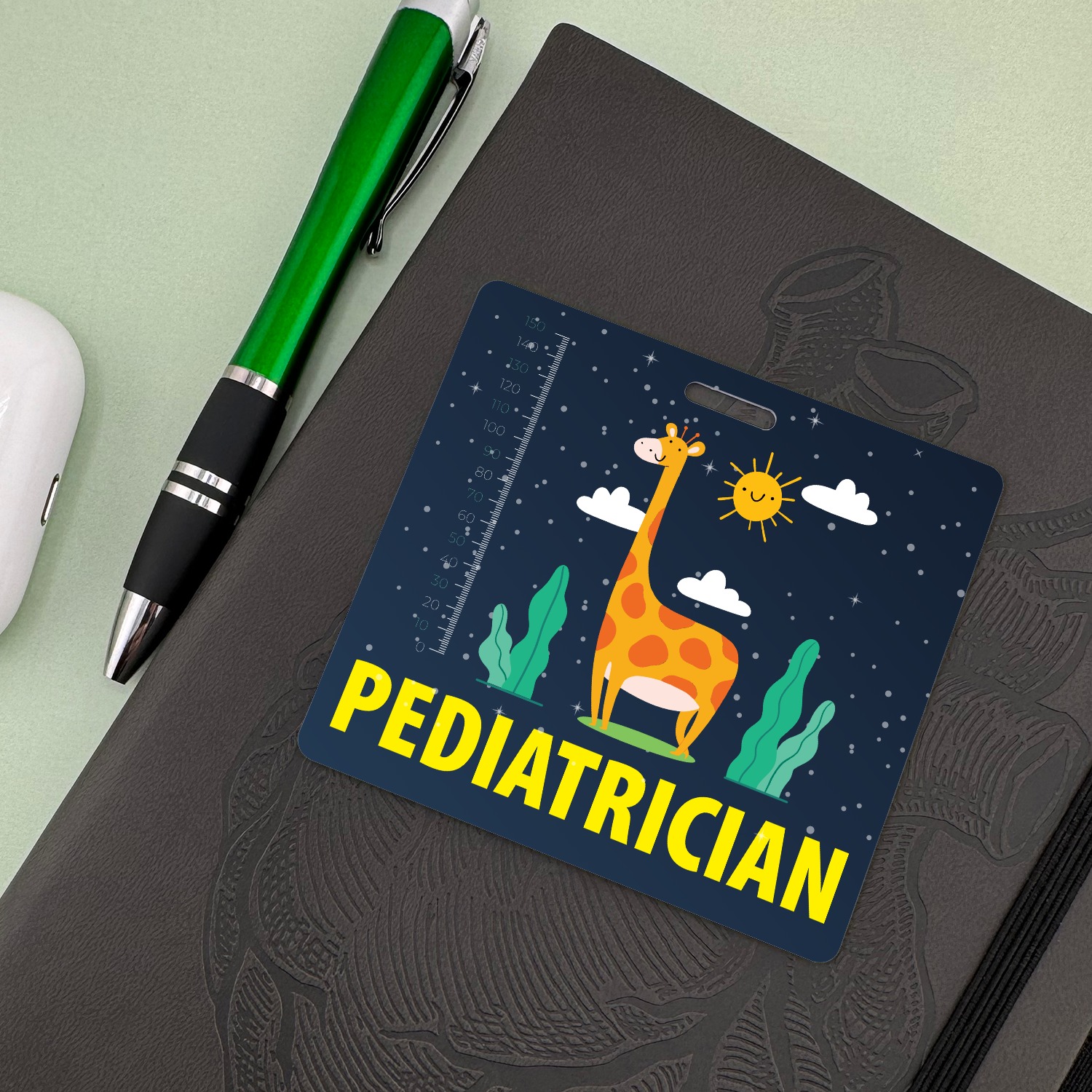 Pediatrician badge buddy with giraffe night sky design beside a green ballpoint pen and white earbud case on a notebook