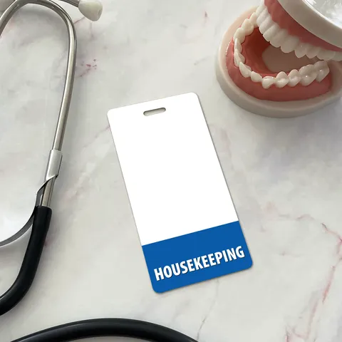 Vertical white housekeeping badge buddy with blue HOUSEKEEPING band on marble surface beside stethoscope and dentures