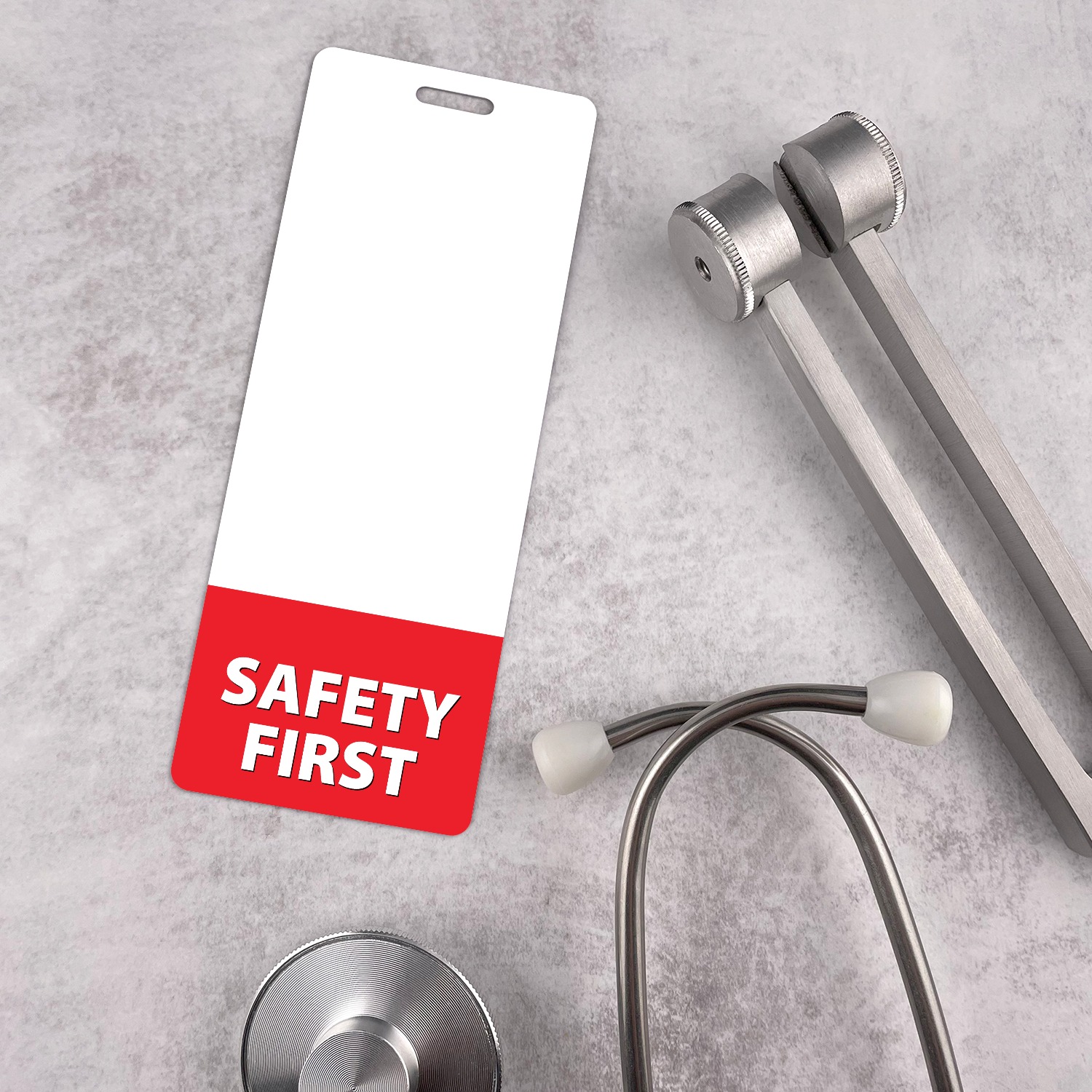 Slim SAFETY FIRST badge buddy beside a silver stethoscope and tuning fork on grey textured background