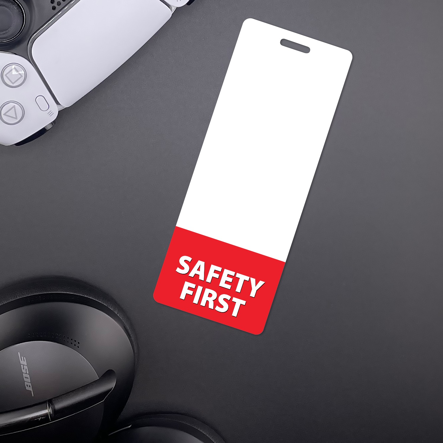 Slim vertical SAFETY FIRST badge buddy beside a white game controller and black headphones on dark desk