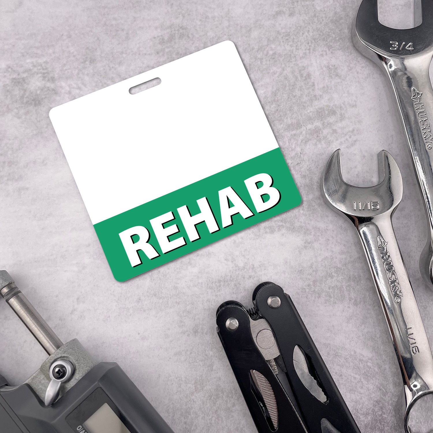 Horizontal REHAB badge buddy with green band among chrome wrenches and multitool on grey surface