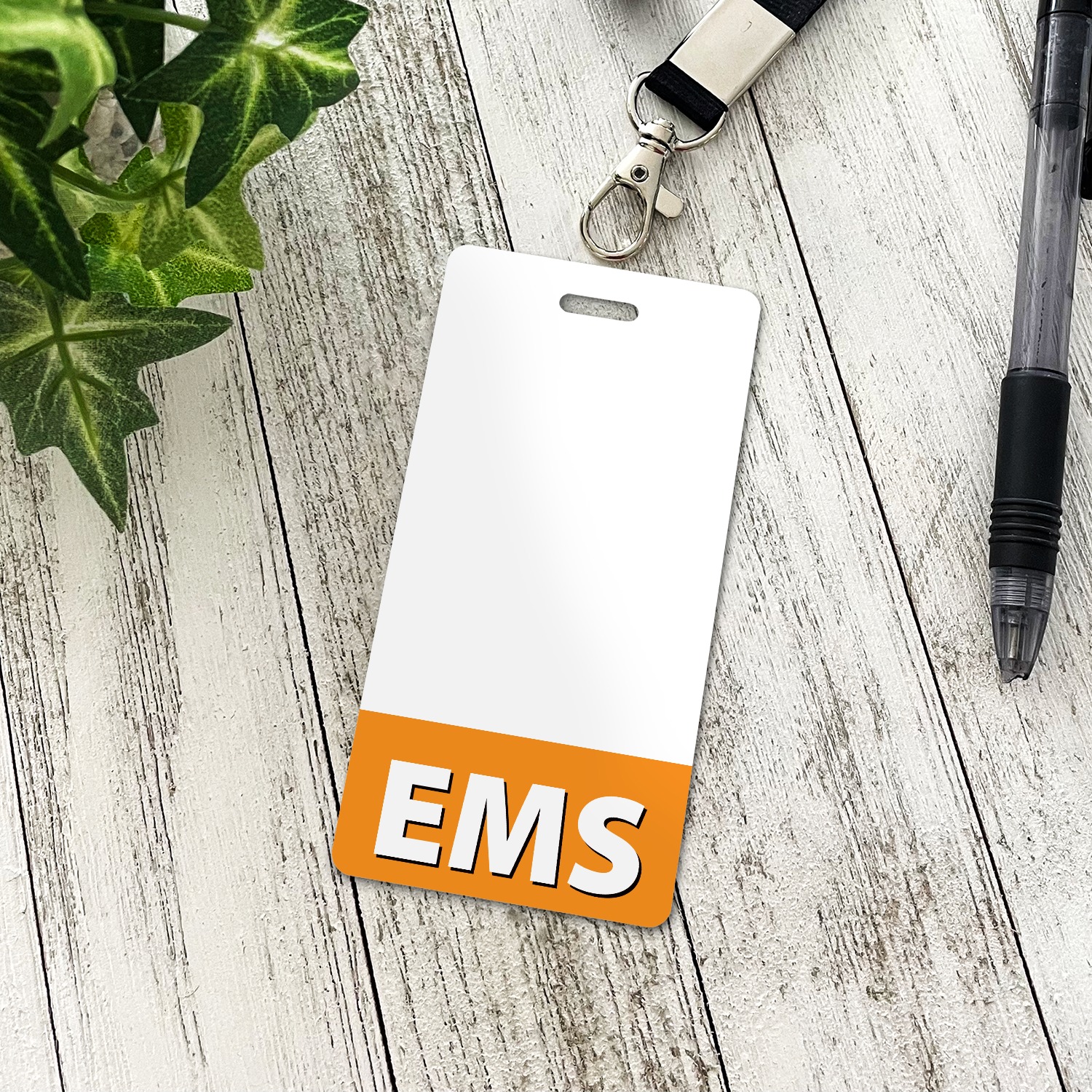 Vertical white EMS badge buddy with orange band and lanyard clip on white wood boards beside a plant and pen