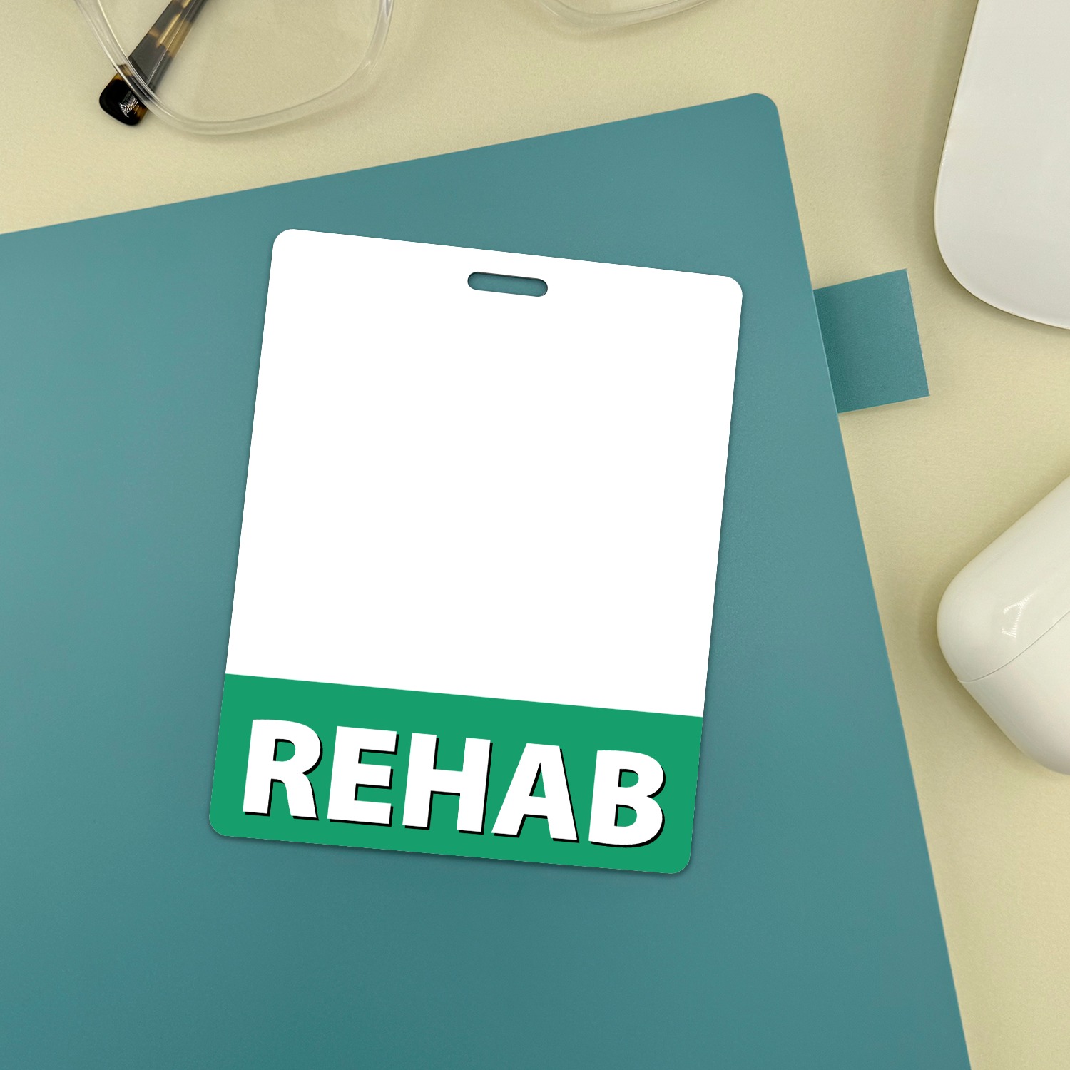 Vertical REHAB badge buddy with green band on teal notebook beside glasses and white mouse
