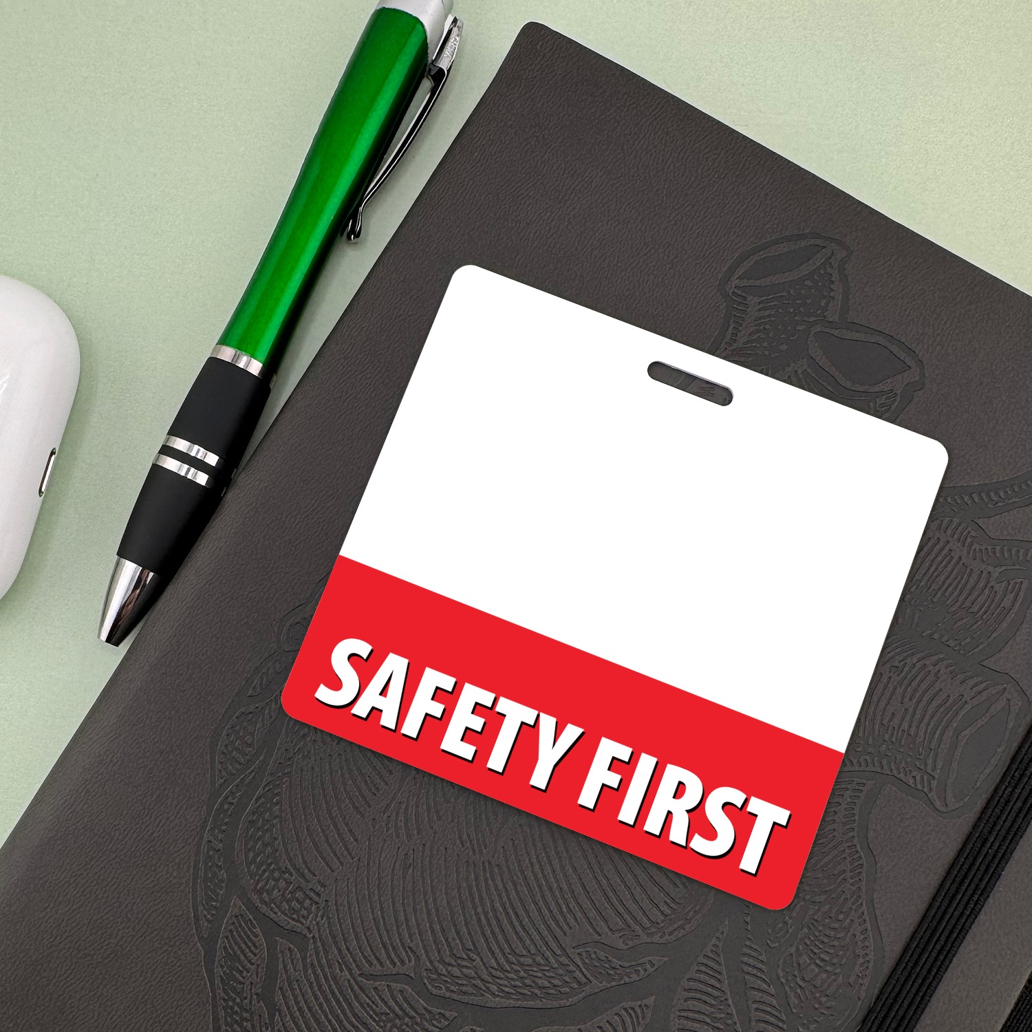 Horizontal SAFETY FIRST badge buddy on a black notebook beside a green ballpoint pen on mint background