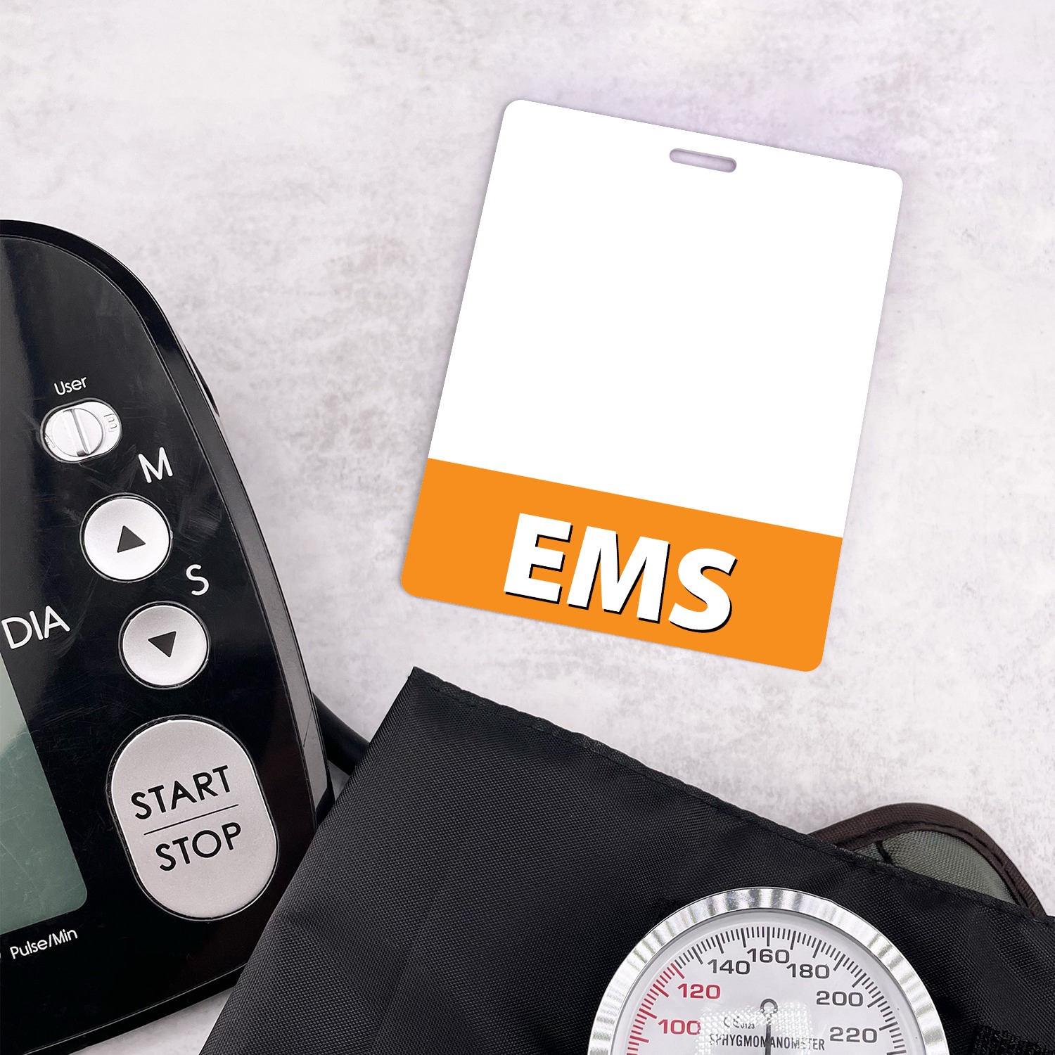 White EMS badge buddy with orange band beside a black blood pressure monitor and silver gauge on marble