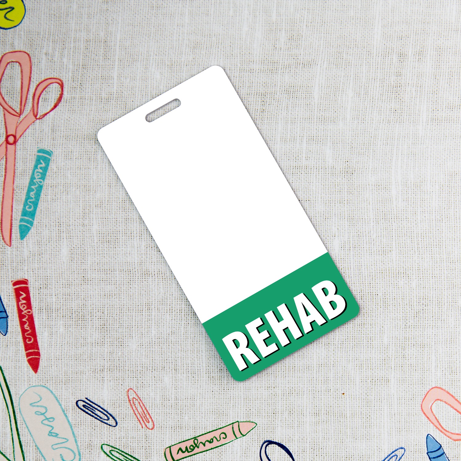 Vertical REHAB badge buddy with green band on grey linen surrounded by colourful kids craft supplies