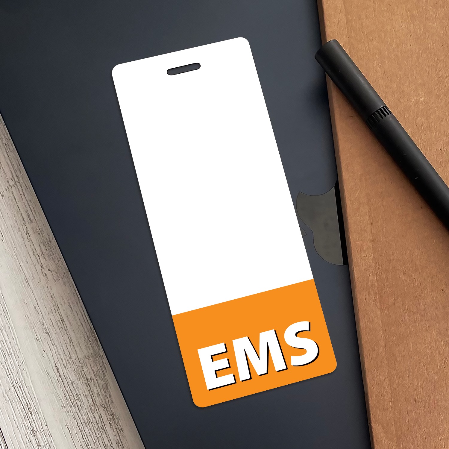 Narrow vertical white EMS badge buddy with orange band on dark grey folder beside a black pen and kraft paper