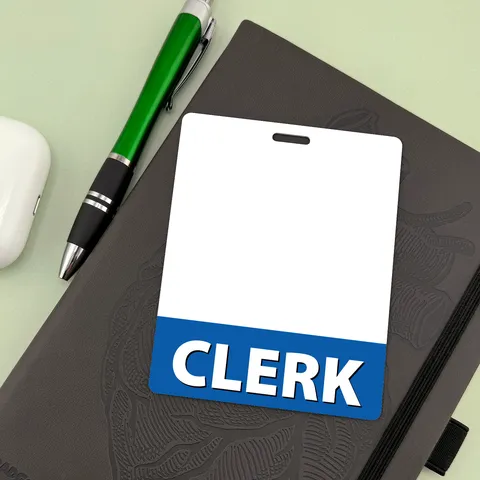 Upright CLERK badge buddy on a dark notebook beside a green ballpoint pen and a white earbud case