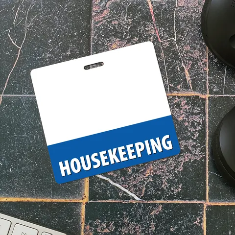 Tilted horizontal housekeeping badge buddy on dark stone tiles with a blue HOUSEKEEPING band beside a keyboard