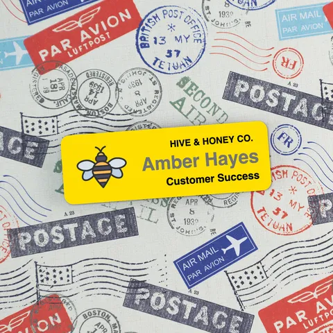 Yellow bee name badge reading Amber Hayes, Customer Success, placed over vintage airmail postage stamp fabric