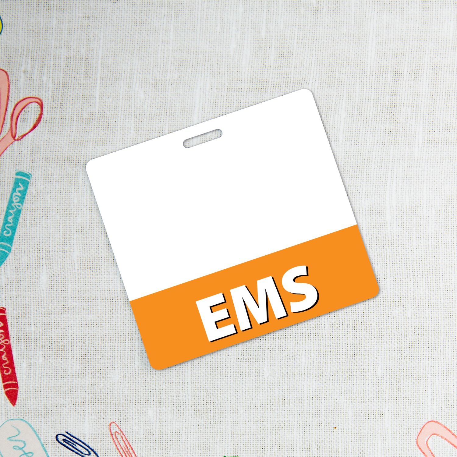 Horizontal white EMS badge holder with orange bottom band on textured fabric beside crayon and scissors doodles