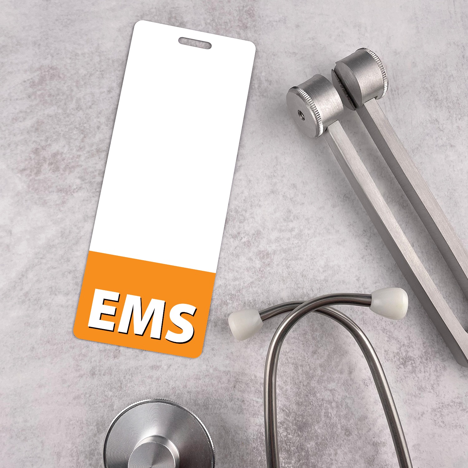 Narrow white EMS badge buddy with orange band beside a silver stethoscope and tuning fork on grey textured surface