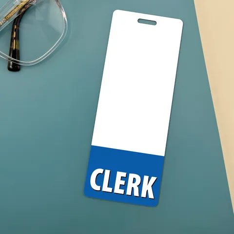 Narrow CLERK badge buddy with blue band on teal desk beside clear eyeglasses with tortoiseshell arms