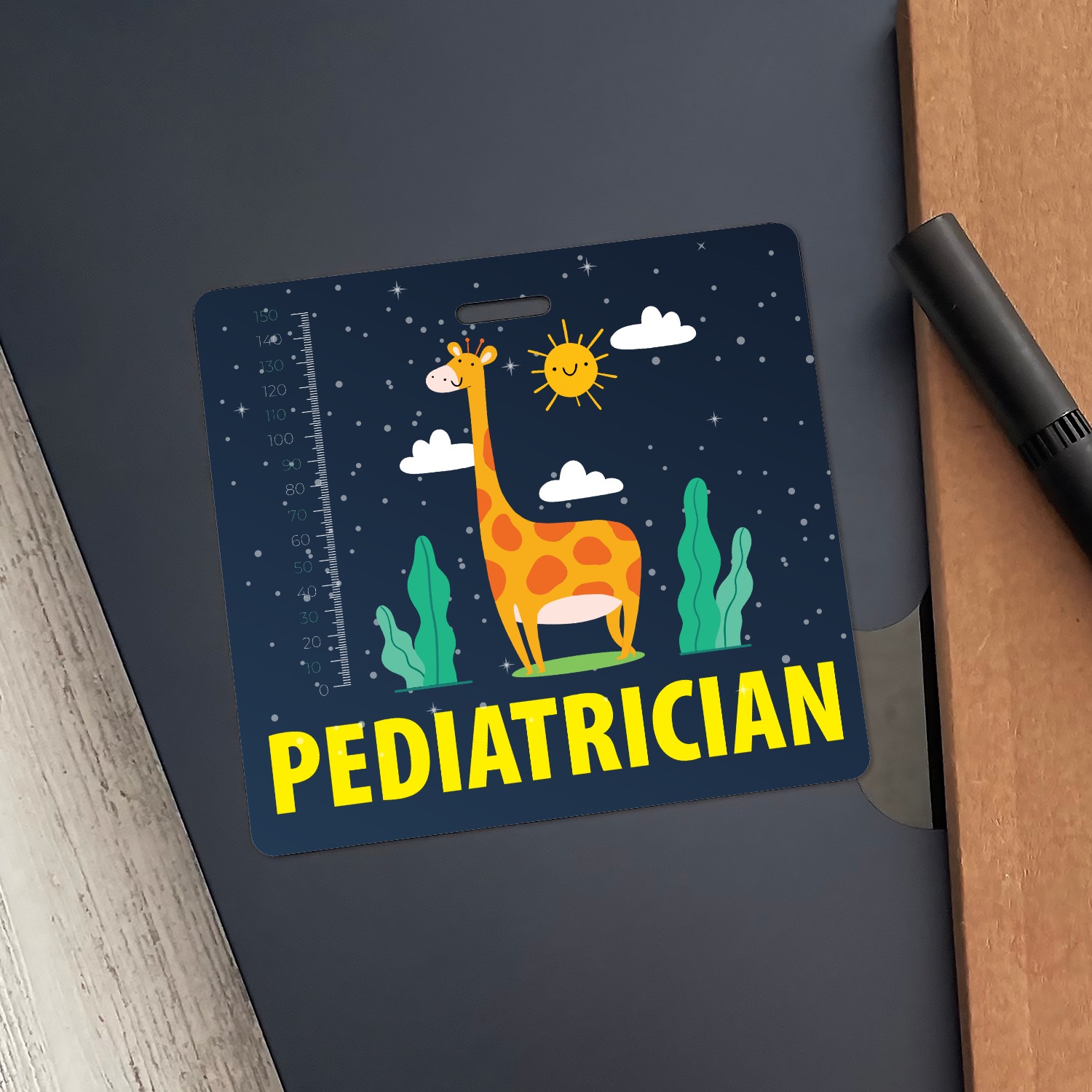 Pediatrician badge buddy with smiling giraffe and height ruler resting on a dark grey folder beside a black pen