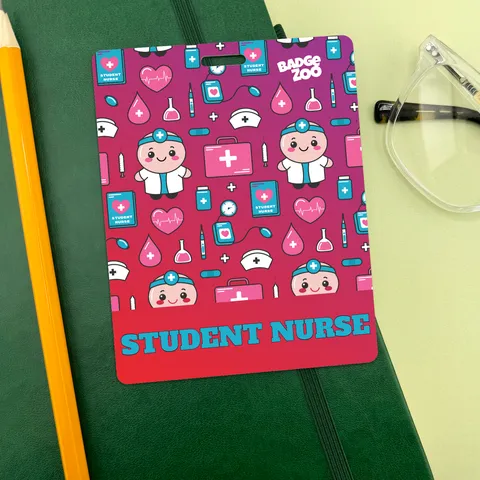 Pink and magenta student nurse ID badge with cartoon nurse characters and medical icons, resting on green notebook beside a