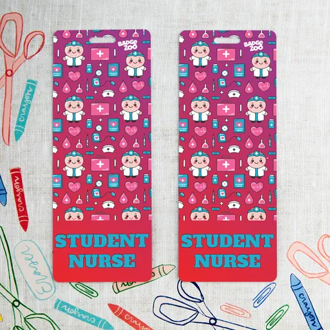 Two student nurse badge buddies with pink medical pattern and teal text, on a table among colorful craft scissors and crayons