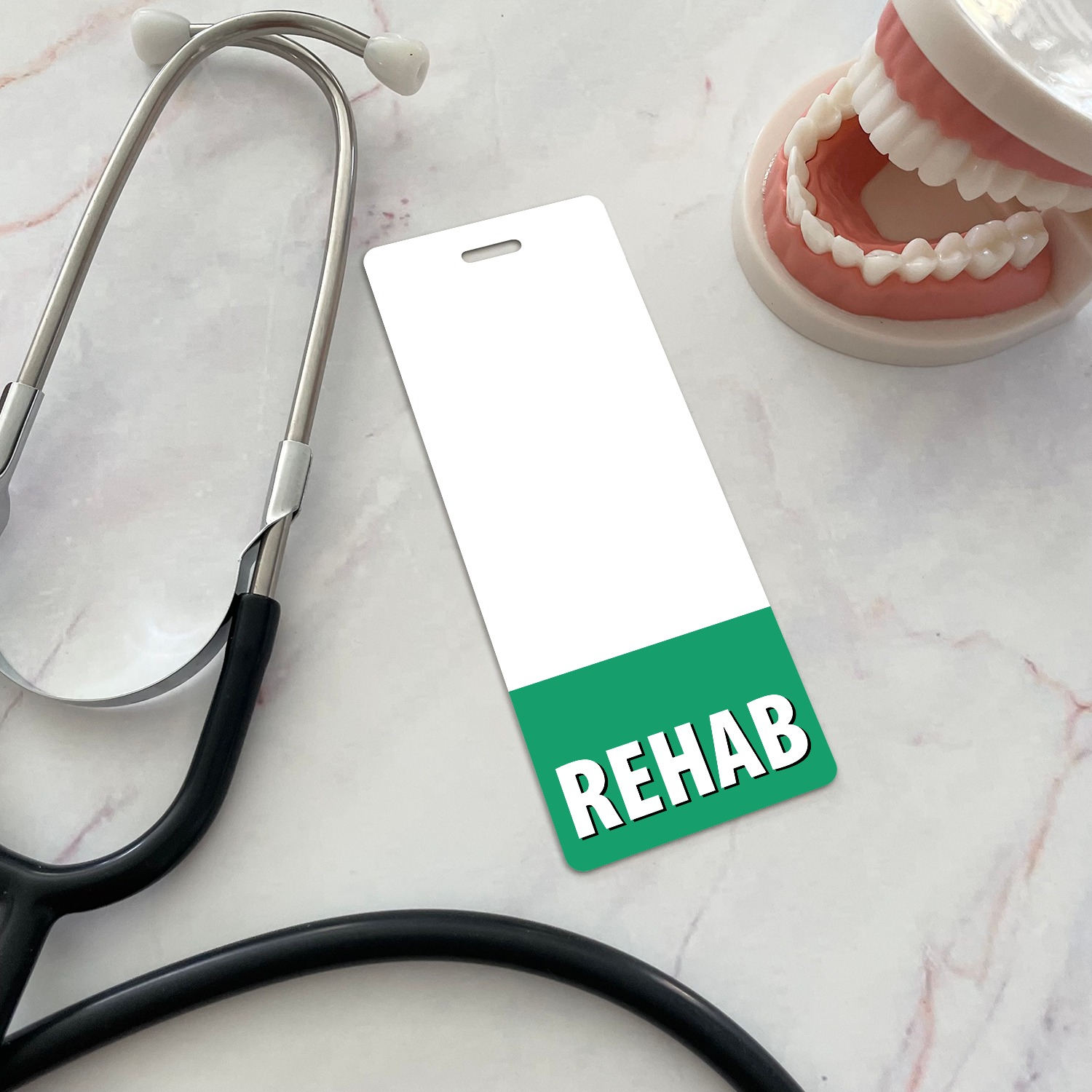 Narrow vertical REHAB badge buddy with green band beside dental jaw model and stethoscope on marble