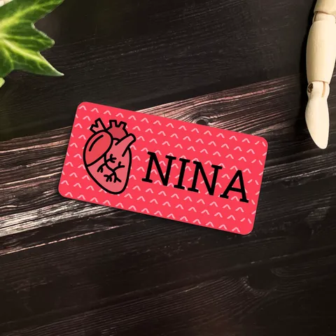 Red heart name badge reading NINA on a dark wood table beside a wooden figure and green ivy