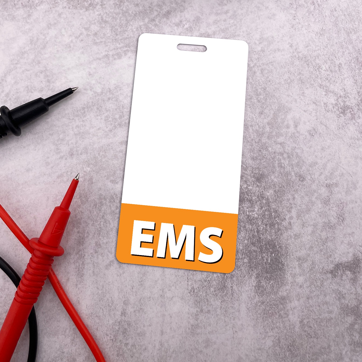 Vertical white EMS badge buddy with orange band on grey surface next to red and black multimeter test leads