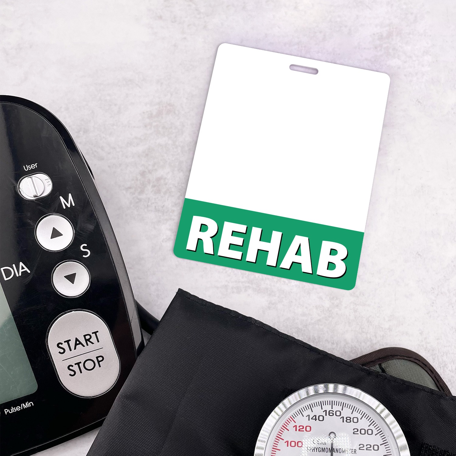 Vertical REHAB badge buddy with green band beside black blood pressure monitor and cuff gauge