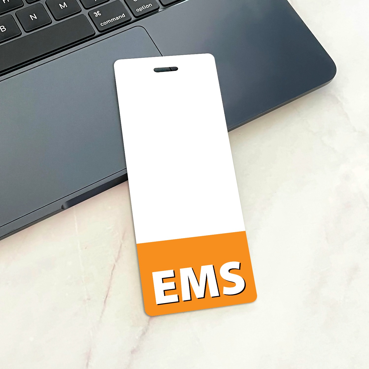Narrow white EMS badge buddy with orange bottom band on dark laptop trackpad beside keyboard on marble surface