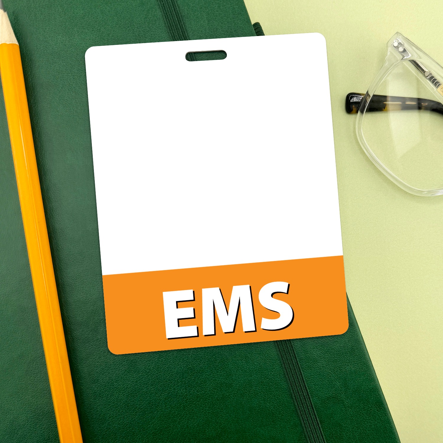 White EMS badge buddy with orange bottom band on green notebook beside a yellow pencil and clear safety glasses