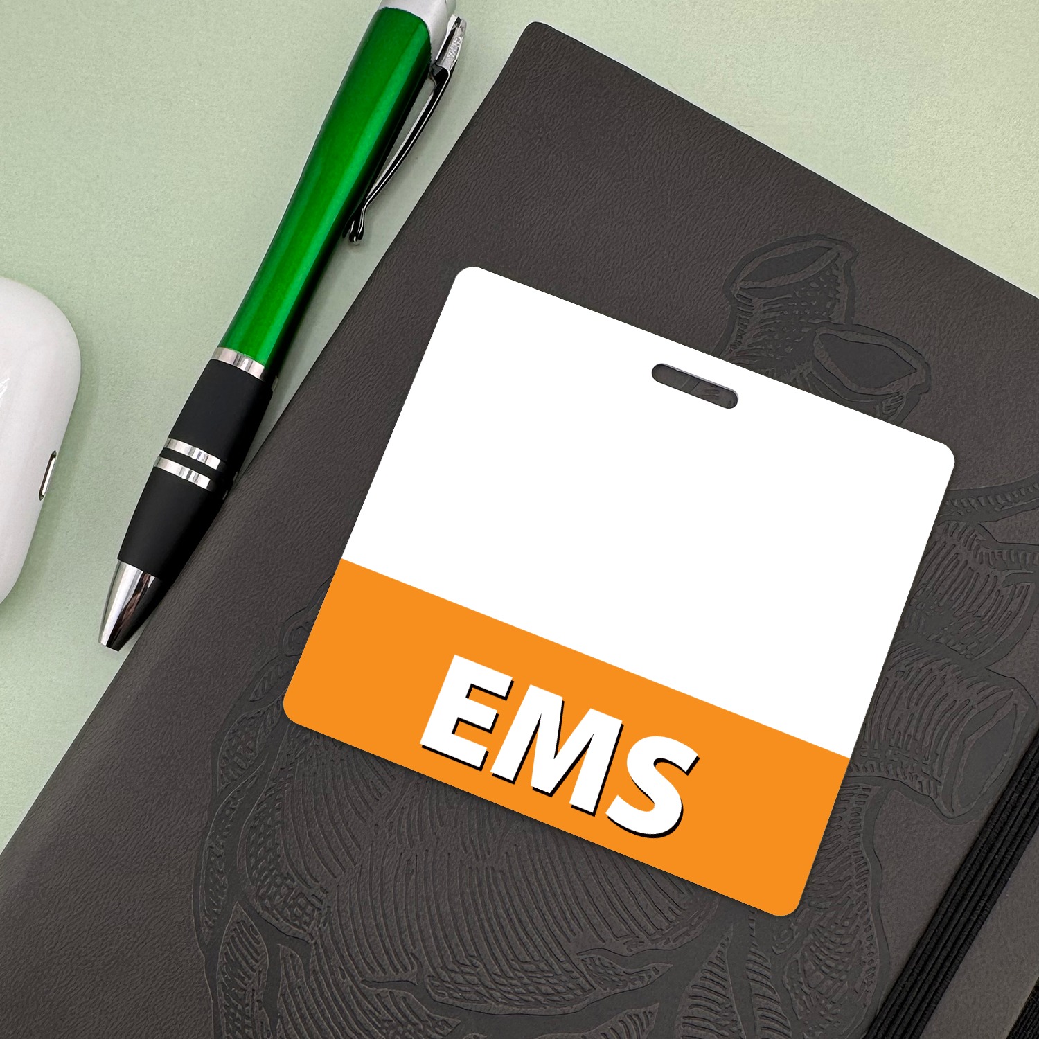 Horizontal EMS badge buddy with orange band on dark notebook beside a green ballpoint pen and white earbuds case