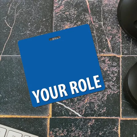 White badge buddy with a top slot and YOUR ROLE text lying on dark tile with a keyboard and mouse nearby.