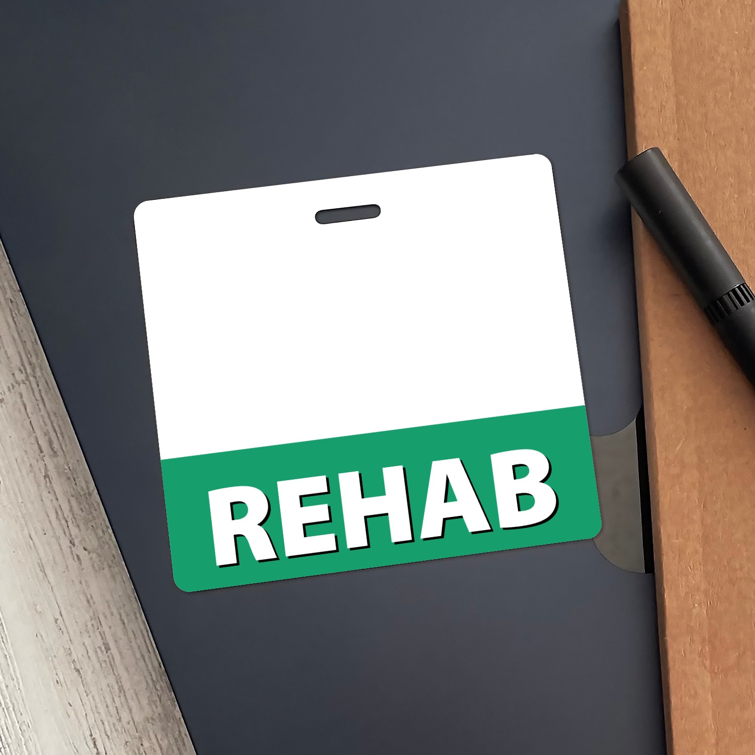 Horizontal REHAB badge buddy with green bottom band lying on dark navy folder beside a black pen and wood