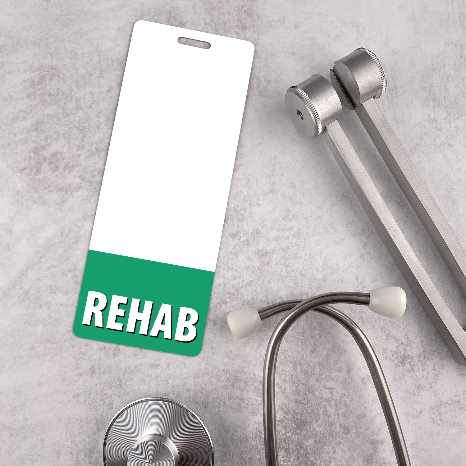 Slim vertical REHAB badge buddy with green band beside black stethoscope on white marble surface