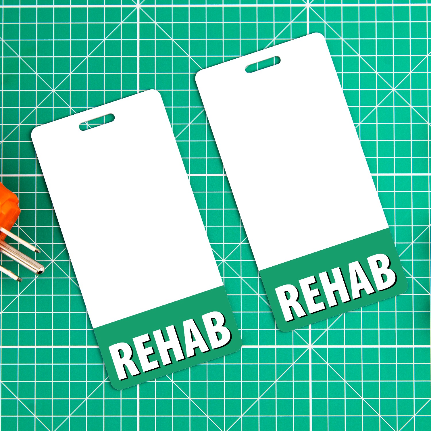 Two vertical REHAB badge buddies with green bottom bands on green cutting mat with small tools