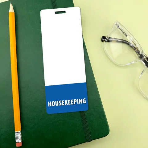 Narrow housekeeping badge buddy with blue HOUSEKEEPING band beside yellow pencil and eyeglasses on green notebook