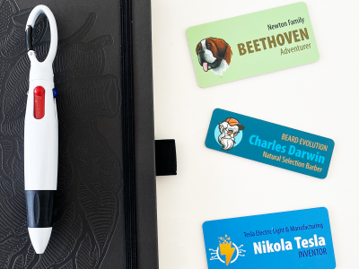 Three custom name tags for pets and professionals next to a black notebook and multi-color pen